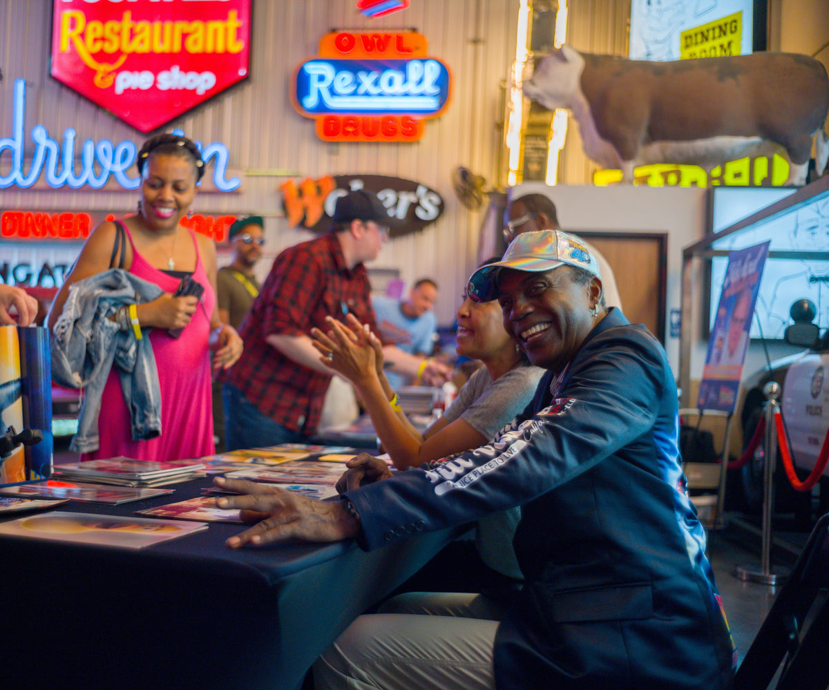 People interacting in a lively indoor setting with neon signs and a vintage car. Don Fullilove smiling sitting at his signing table during the 40th Fan Bash in Los Angeles CA hosted by Doc's Time Machines. 